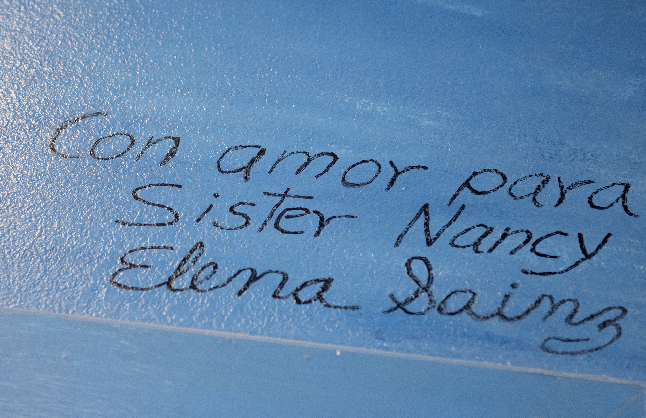 The words “Con Amor Para Sister Nancy,” is written in cursive with black paint on a blue wall. The translation of the words is, “with love for Sister Nancy.”