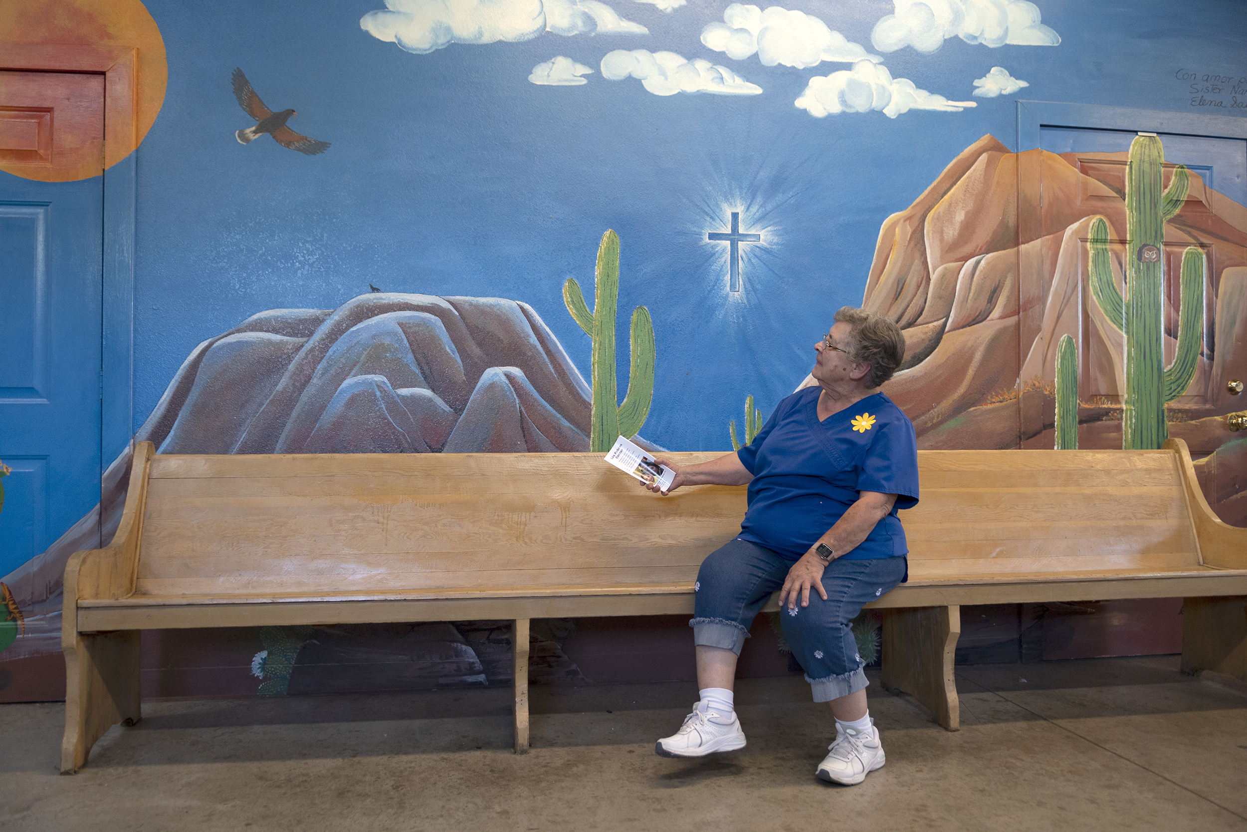 Sister Nancy sits on a donated pew bench and looks up at artwork behind her depicting a white cross centered within a bright blue mural depicting mountains, a cactus, hawk, and the sky.
