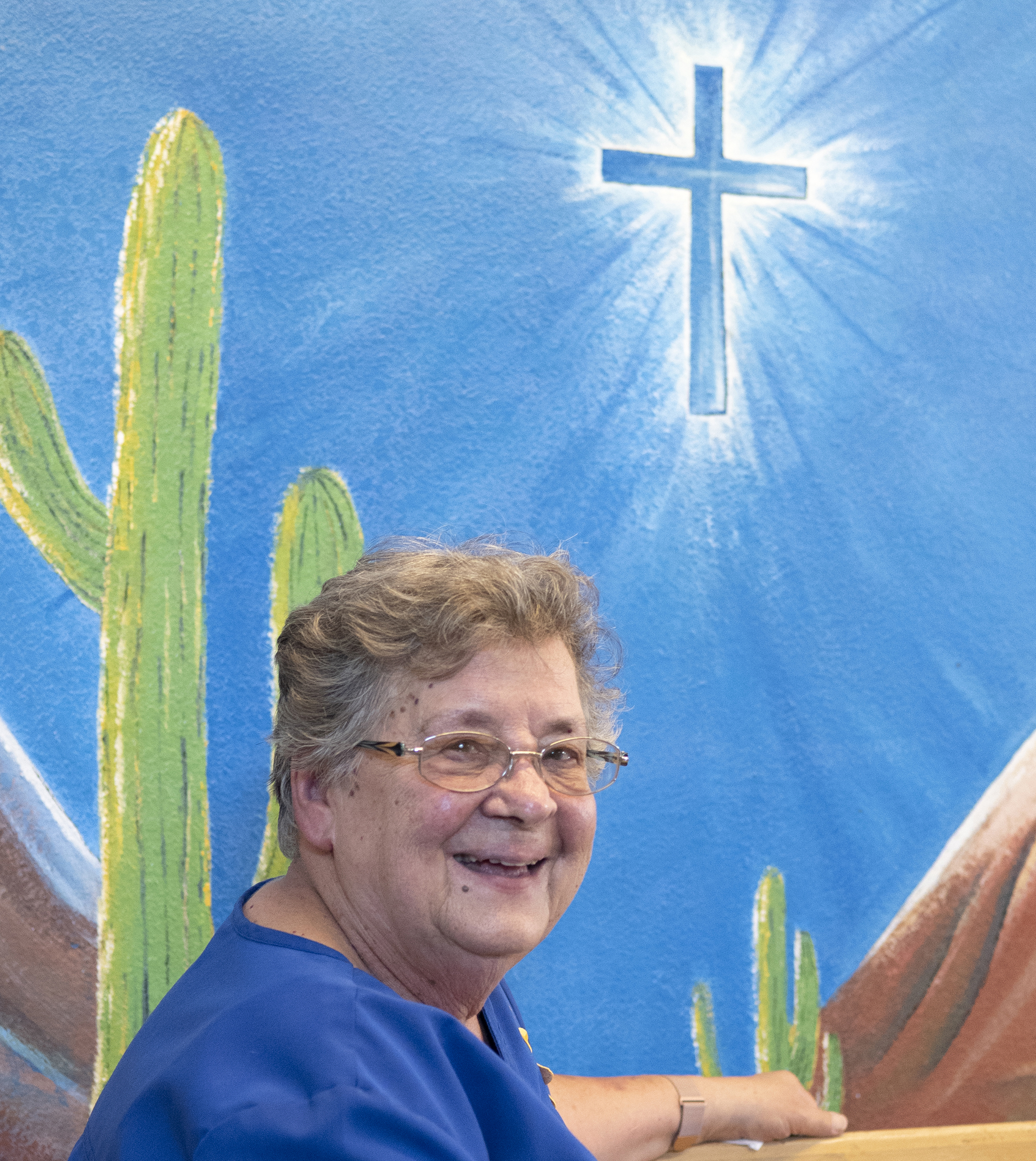 Sister Nancy has round-rimmed glasses and short salt and pepper hair. She smiles while wearing a blue top and sits under a wall mural with a green cactus and white cross.
